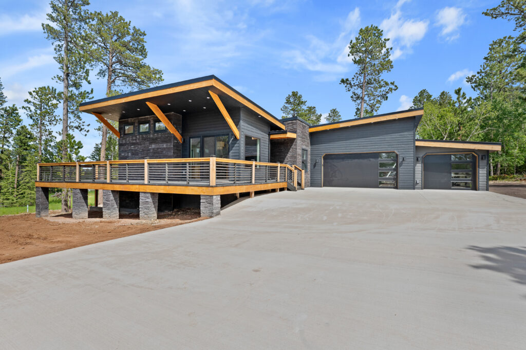 Mead Lumber's Legace house plan, Beethoven, built in Lead, SD. Gray sided 1-story house with 2 car garage with angled roof and front elevation with deck, gray foundation pillars, and wood beam accents.