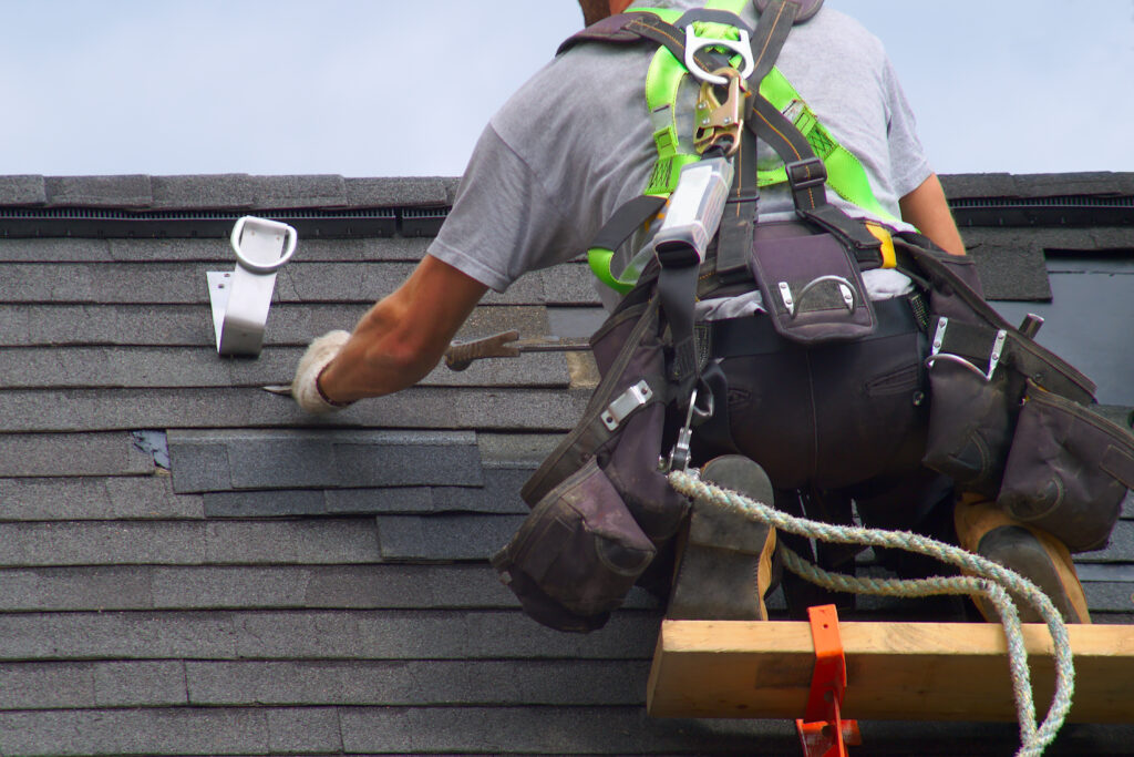 The back of a roof installer kneeled on a wood board, wearing a safety harness and work boots, has a hammer and a small cup on nails, on a gray shingles roof.