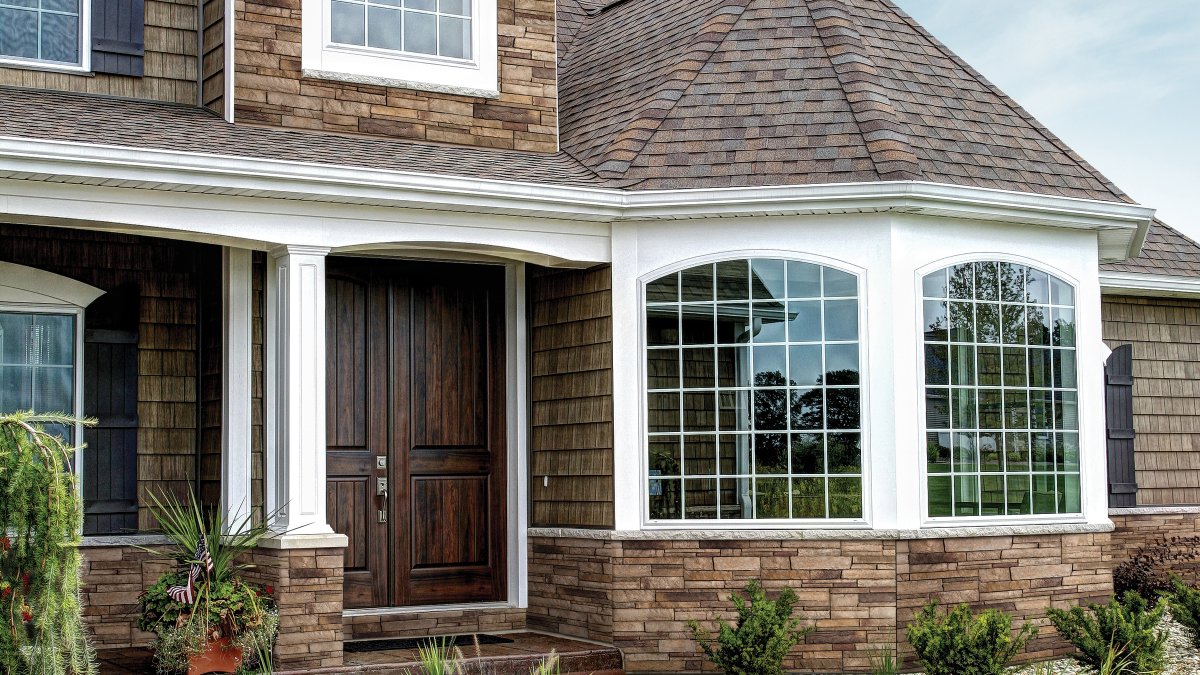 House with double wood front doors, brown roof shingles, brown stone front and brown shake siding, white columns, and 2 picture windows.