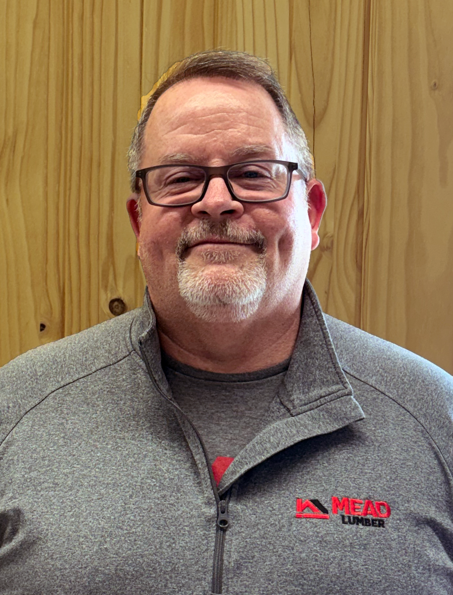 Allan Wilson headshot with short brown hair, glasses, a gray zip polo with a red and black Mead Lumber logo on the chest and a light colored wood in the background.