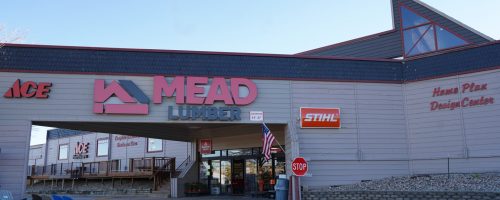 An exterior shot of the Mead Lumber of Rapid City, SD West store with an roof extended over the doors with brown siding and black scalloped shingles above and Mead Lumber, Ace, Stihl logo signs, underneath there's a US flag hanging and blue Adirondack chairs for sale, a deck railing display and a stop sign